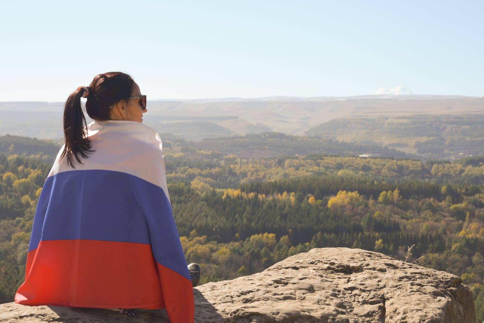 A girl tourist with a Russian flag on her shoulders sits on the top of a mountain and enjoys the beautiful views of the Caucasus Mountains