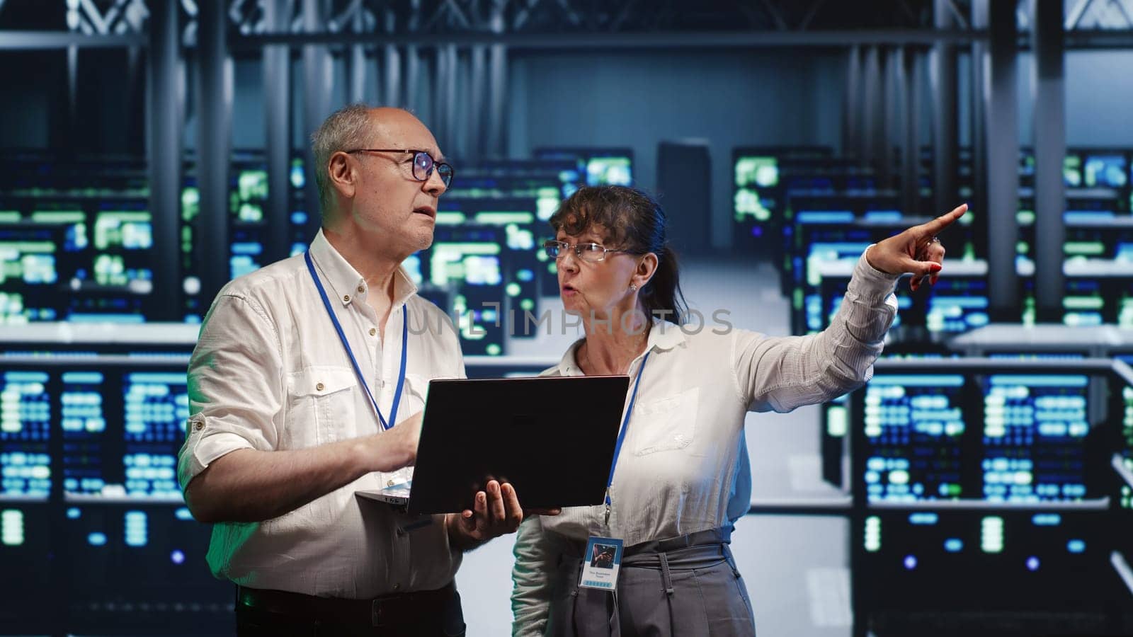 Proficient repairmen looking around high tech data center, using laptop to crosscheck disaster recovery plan and assess server rows in need of replacement, preventing hindrances