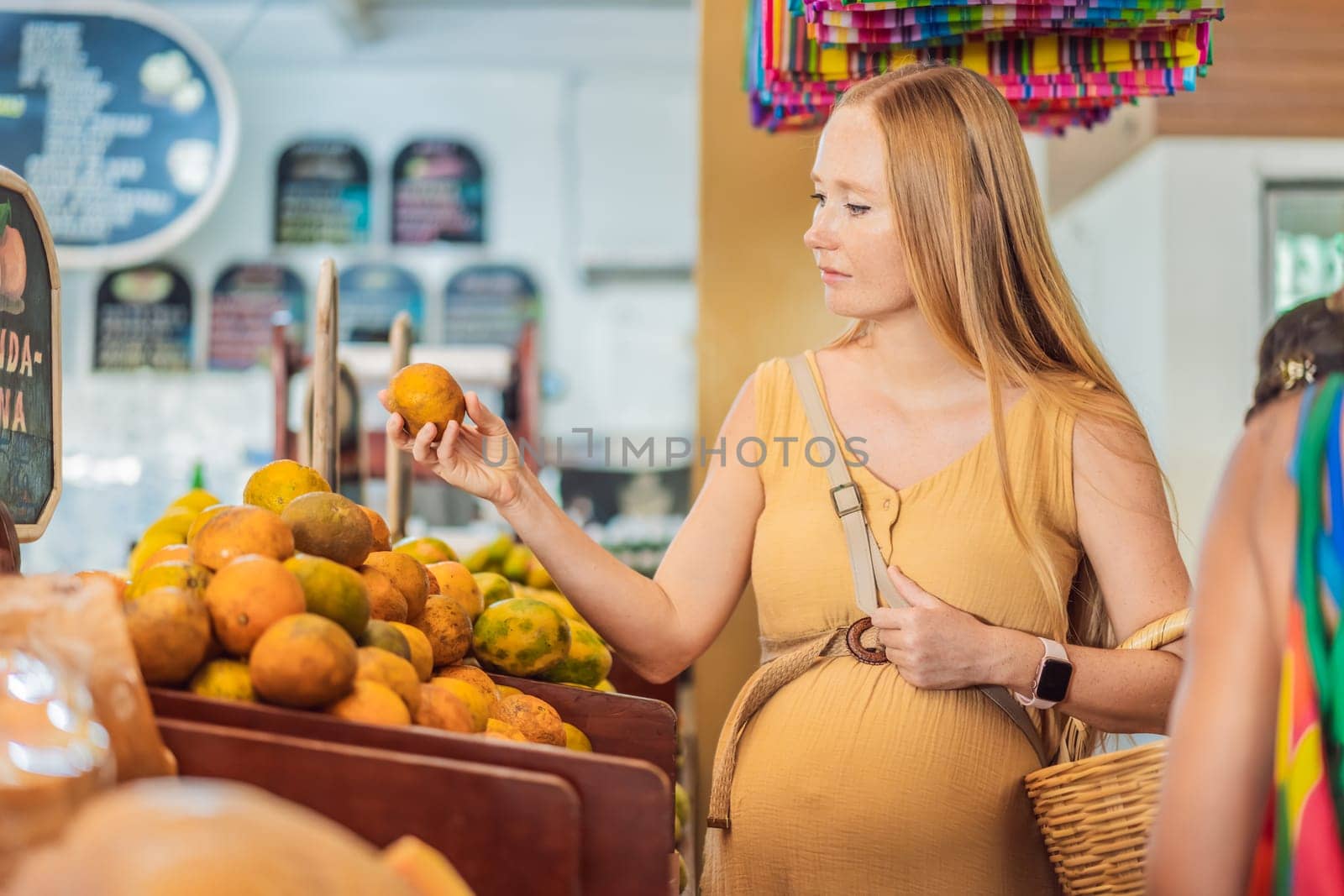 In a grocery store, a pregnant woman stands by a fruit stand, surrounded by various natural foods. She is in a public space where the local market offers whole foods for trade Pregnant woman buying organic vegetables and fruits at Mexican style farmers market