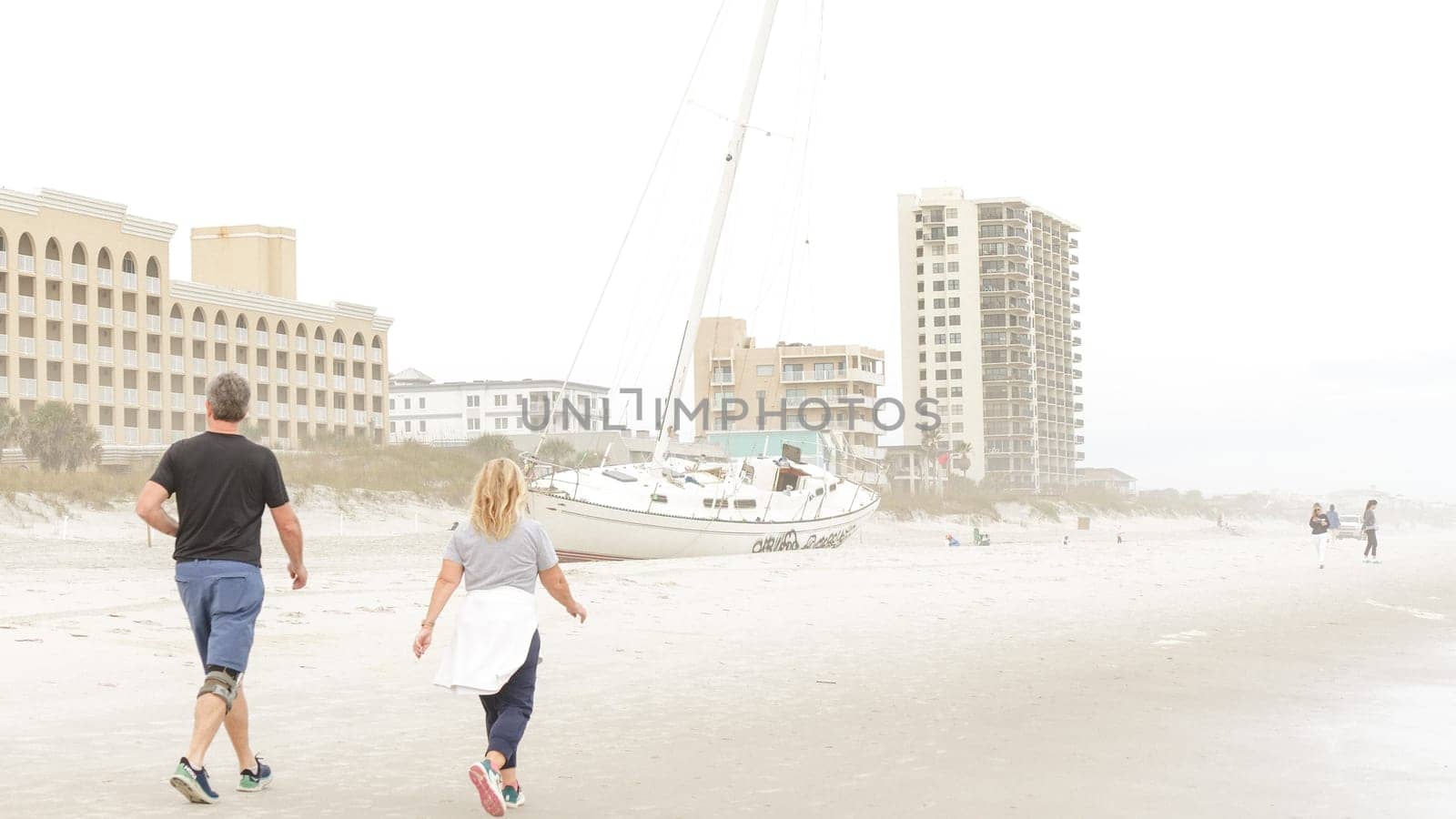 A man and woman are walking away from a beached sailboat on a foggy day. The image is taken from a low angle, making the boat look larger than life. There are buildings in the background.