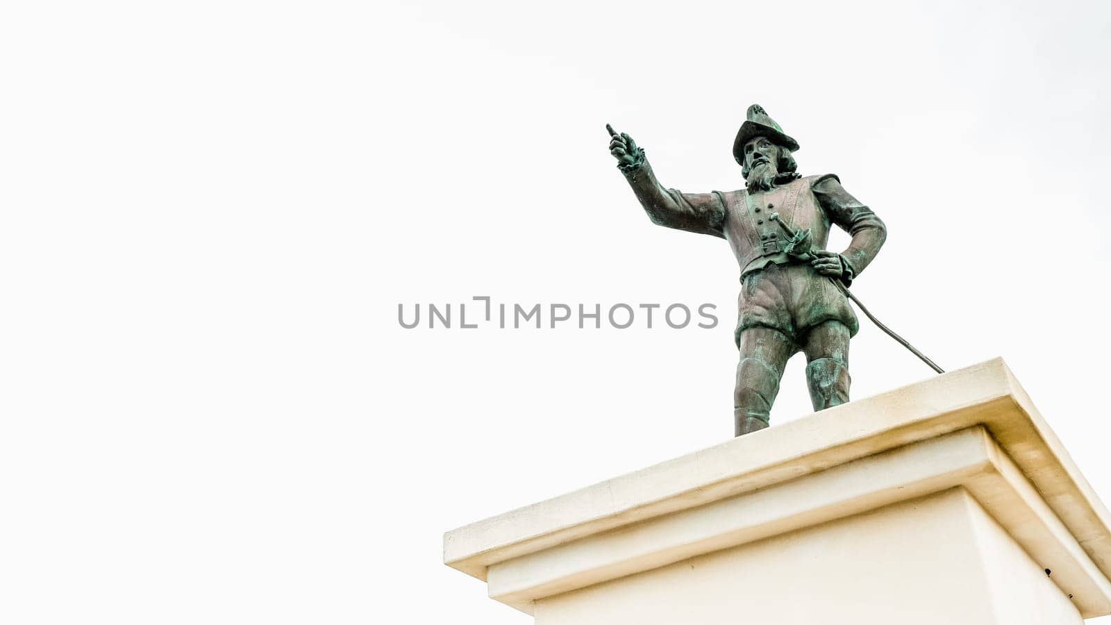 A bronze statue of Christopher Columbus stands atop a marble pedestal, pointing at the horizon with one hand while holding a map in the other. The statue is set against a solid white background.