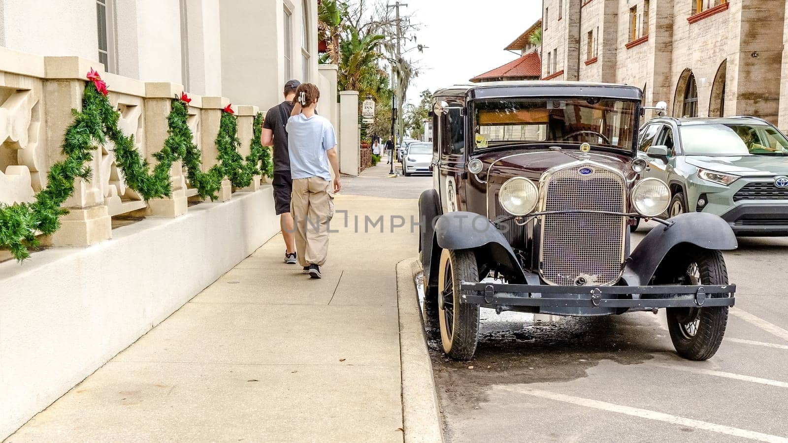 A vintage black car parked on a town road with two pedestrians walking by. The car features shiny black paint, chrome bumpers, and white wall tires, set against buildings and palm trees.
