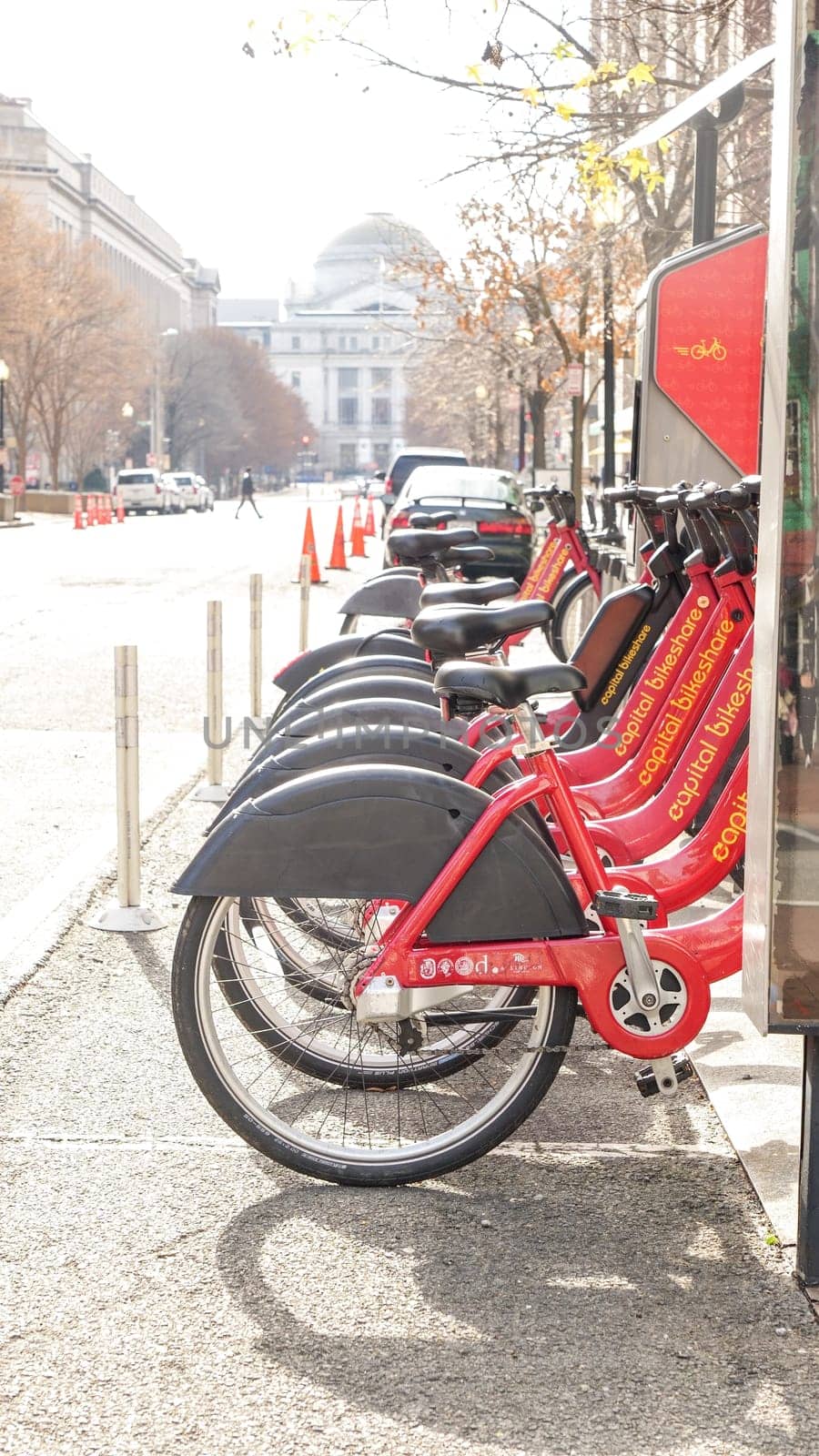 A vibrant row of red bicycles available for rent in the city, lined up near the sidewalk on a sunny day. Background shows blurred cars on the street and a pedestrian walking by.