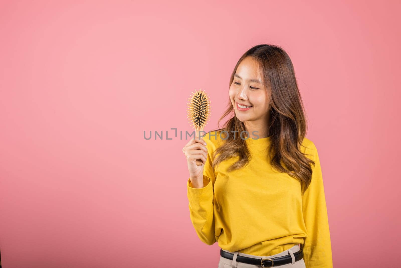 Portrait of Asian beautiful young woman smile combing her hair, happy female long healthy hair holding with hairbrush comb, studio shot isolated on pink background