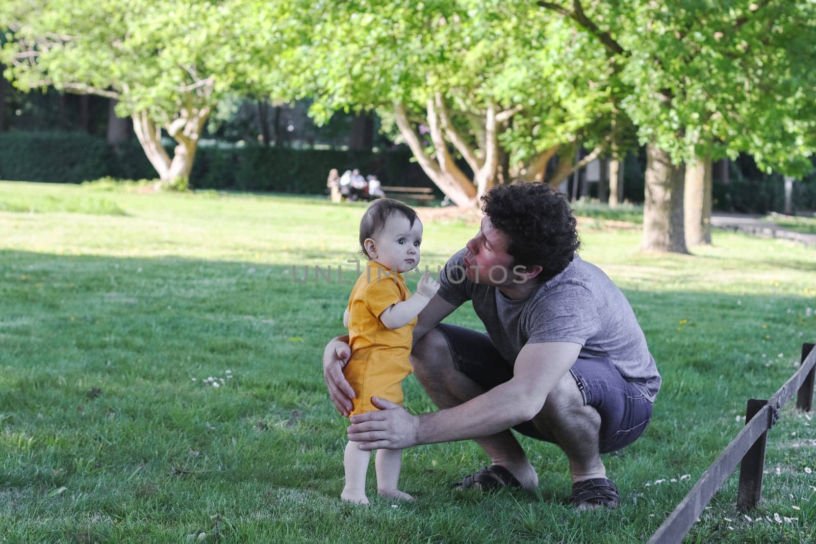 Caucasian young dad in a gray t-shirt, shorts with curly brown hair holds his little daughter in yellow overalls, standing barefoot on the grass, holding a daisy and reaching out to gently kiss her on the cheek in a public park, close-up side view. The concept of fatherhood, dads, family vacation, spring walk, first steps, happy childhood, PARKS and REC.