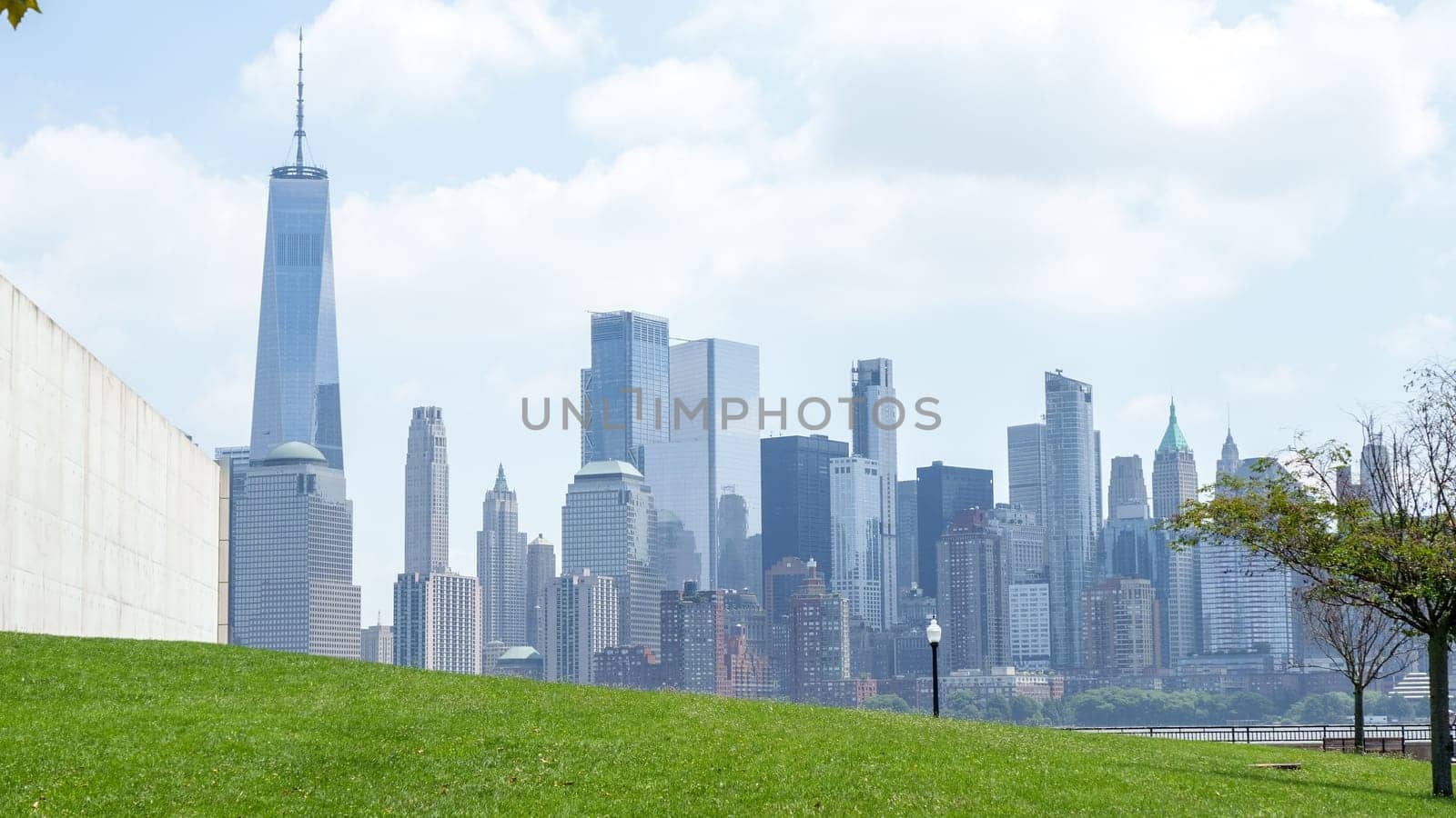 Breathtaking view of Manhattan skyline from Brooklyn Bridge Park on a sunny day. Clear blue sky, white clouds, shining sun, green grass. Skyscrapers, apartment buildings in background.