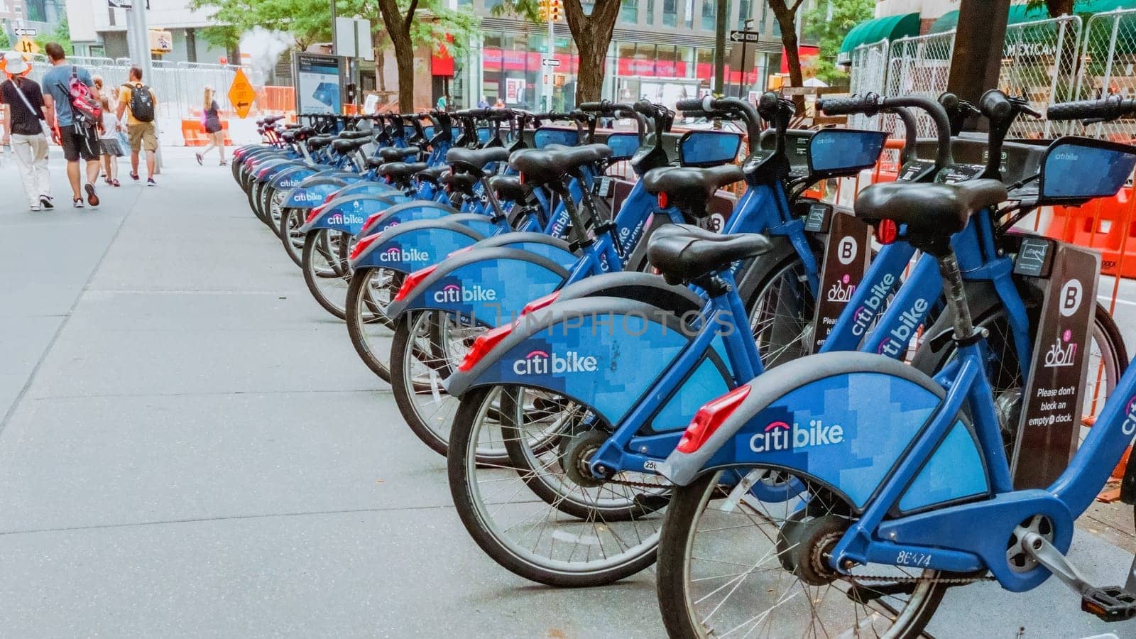 A row of vibrant blue bicycles neatly lined up on a bustling city street. Pedestrians walk by as tall buildings form a picturesque urban backdrop, creating a colorful scene in the city environment.