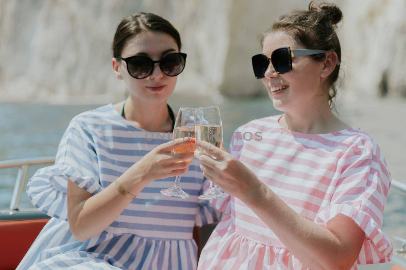 Portrait of one young Caucasian beautiful couple of girls in sunglasses sitting in a boat and knocking glasses of champagne with a happy smile against the backdrop of blurred rocks while sailing on the sea on a sunny summer day, close-up side view.