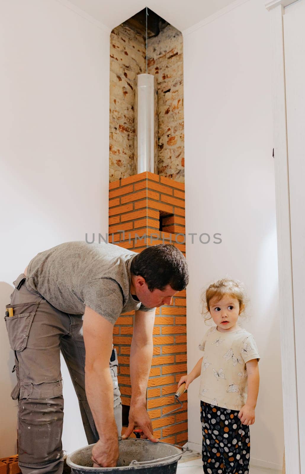 One young Caucasian man in a work uniform stands bent over and kneads cement mortar in a bucket, a small daughter stands nearby and looks to the side while standing in a room where repairs and installation of a smoker for a fireplace made of bricks are underway, close-up side view. The concept is clean brickwork.