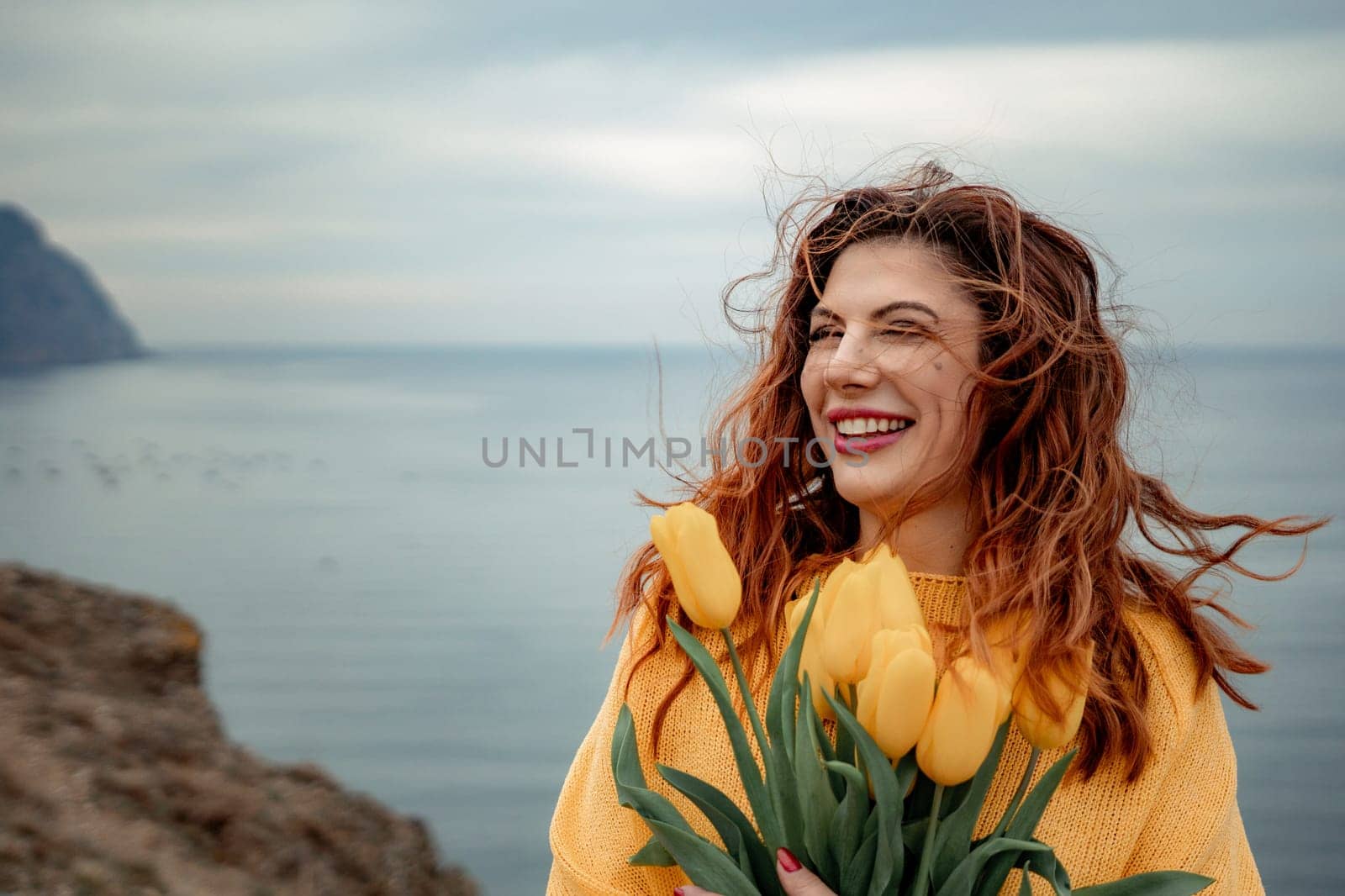 Portrait of a happy woman with hair flying in the wind against the backdrop of mountains and sea. Holding a bouquet of yellow tulips in her hands, wearing a yellow sweater by Matiunina