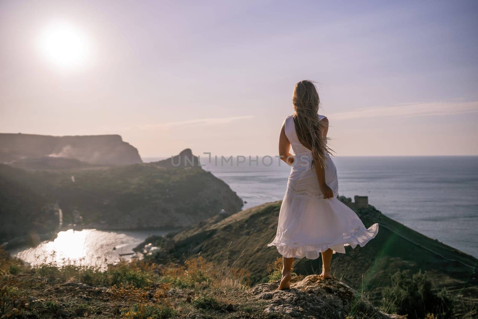 A woman in a white dress stands on a hill overlooking the ocean. The scene is serene and peaceful, with the woman's dress billowing in the wind. The combination of the ocean