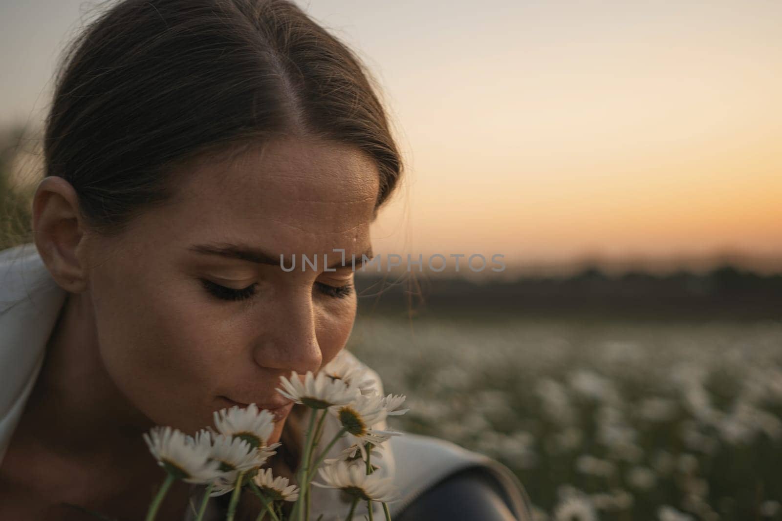 A woman is smelling flowers in a field. The flowers are white and the sky is orange. The woman is looking at the camera