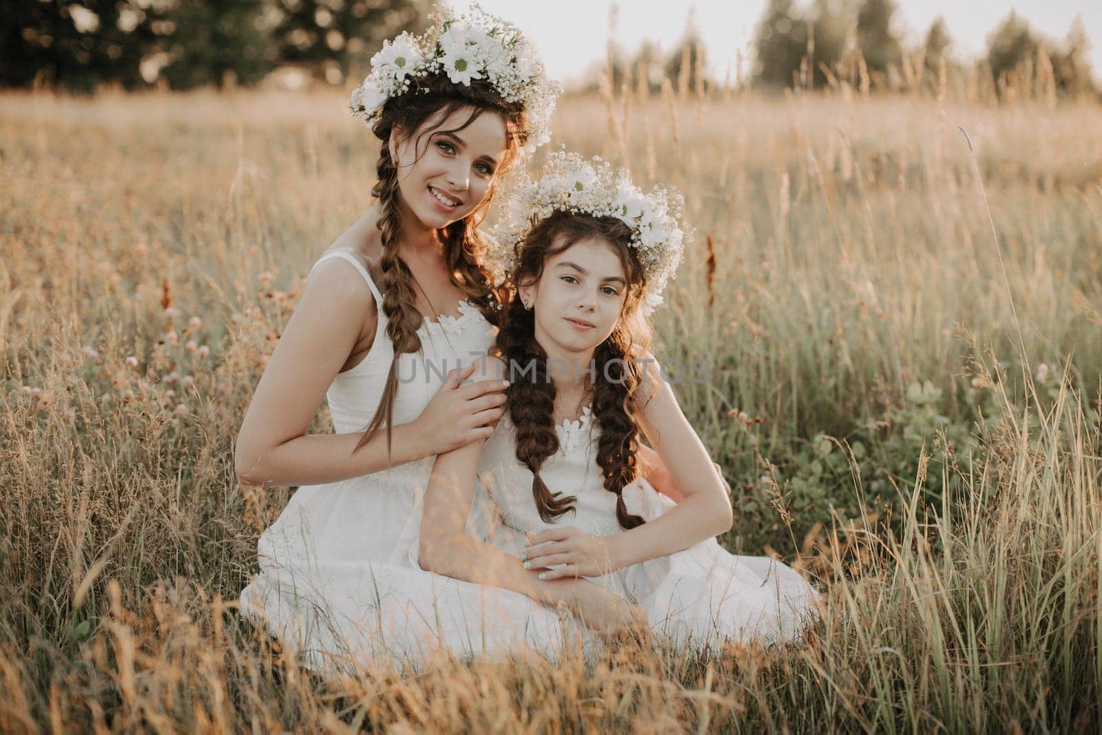 Happy mom and daughter smiling and hugging in field in the summer. Sisters in white dresses with floral wreaths and braids in boho style. Added a small grain simulation of film photography