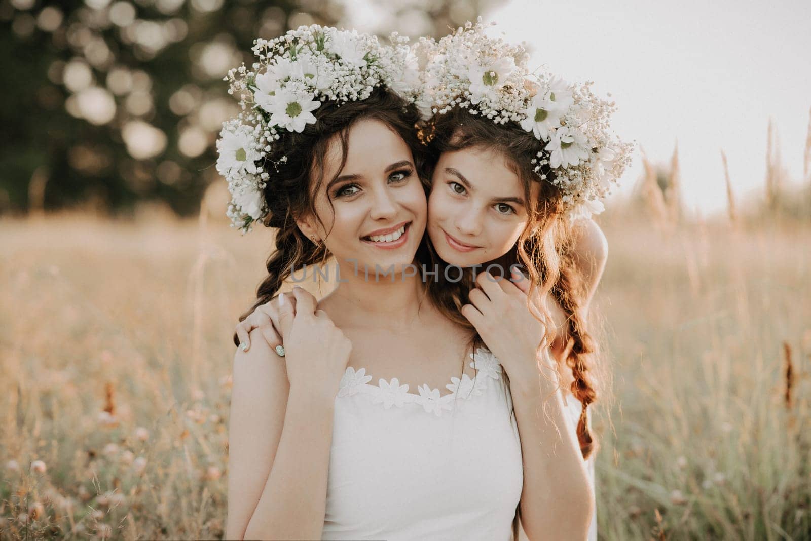 Happy mom and daughter smiling and hugging in field in the summer. Sisters in white dresses with floral wreaths and braids in boho style. Added a small grain simulation of film photography