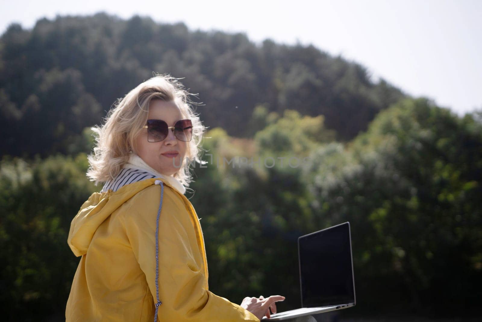 A woman in a yellow jacket is sitting on a bench with a laptop in front of her. She is smiling and she is enjoying her time outdoors