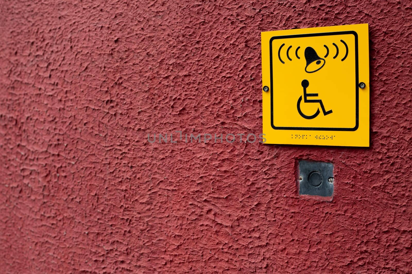 Wheelchair sign with braille text next to staff assistance call button on red structural wall, handicapped access symbol