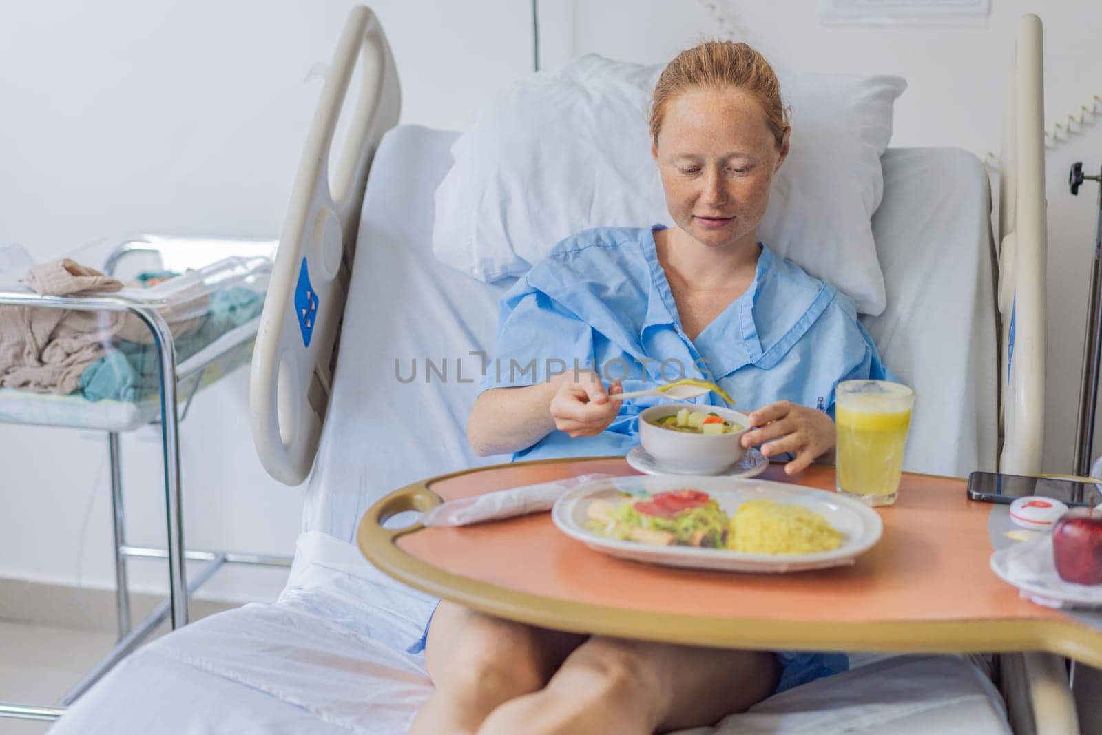 A woman after giving birth in a hospital bed eats her lunch and her baby sleeps in a crib next to her. This moment highlights the early postpartum period, focusing on maternal recovery and the peaceful presence of the newborn.