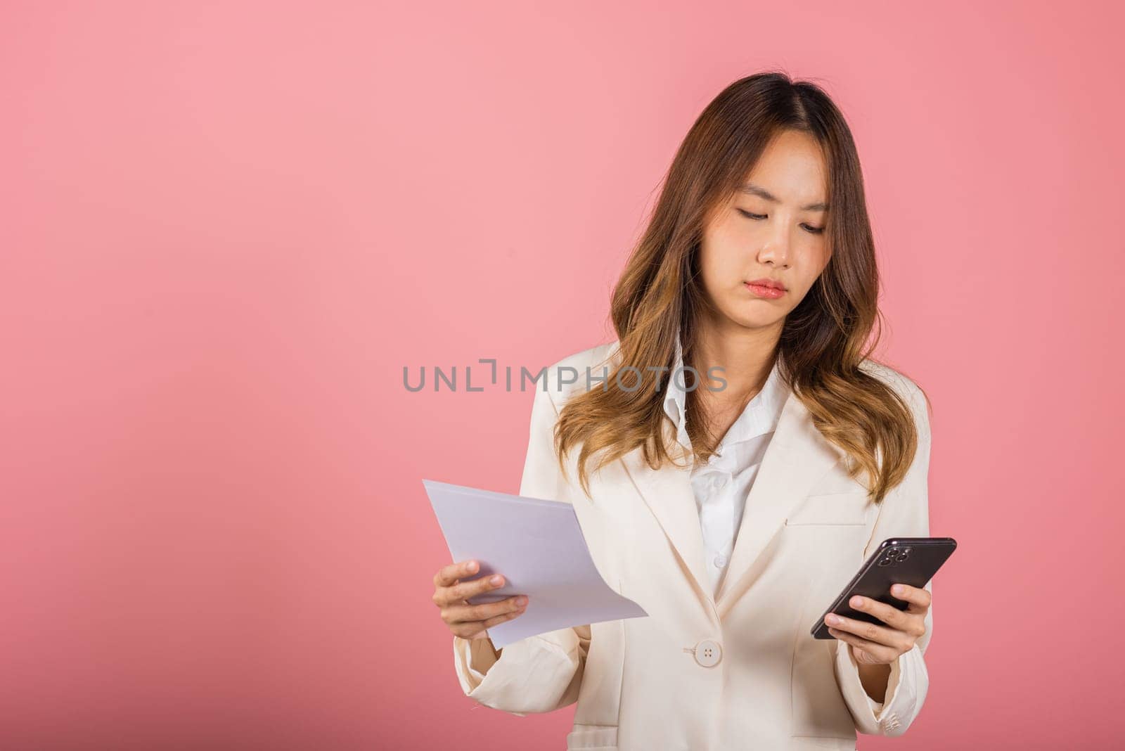 Portrait of Asian young woman holding paper calculating bills on hand and mobile phone for payment QR code, female person problem she hold worried over bill studio shot isolated on pink background