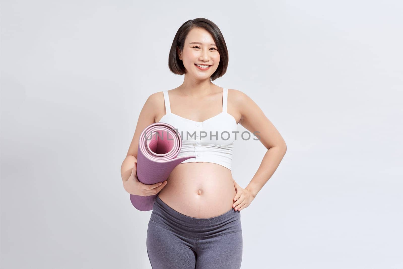 Pregnant girl holding yoga mat standing over white background.