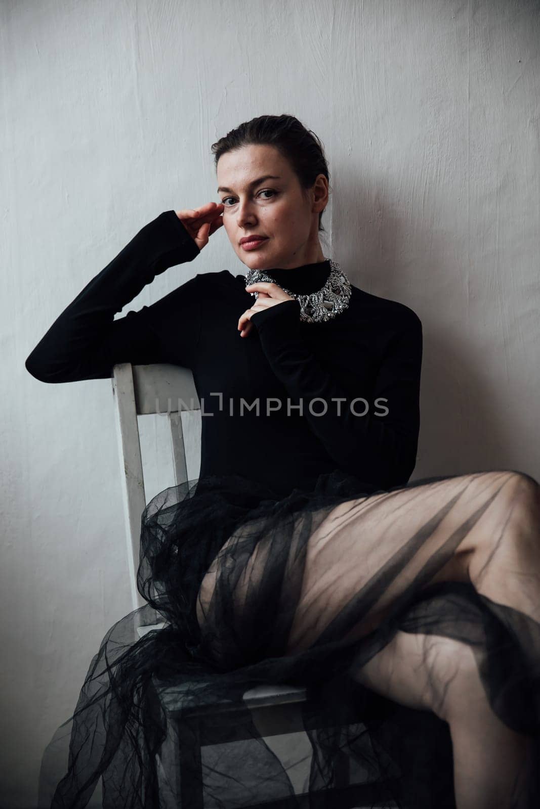a woman in black clothes with tulle sits on a chair against a gray wall