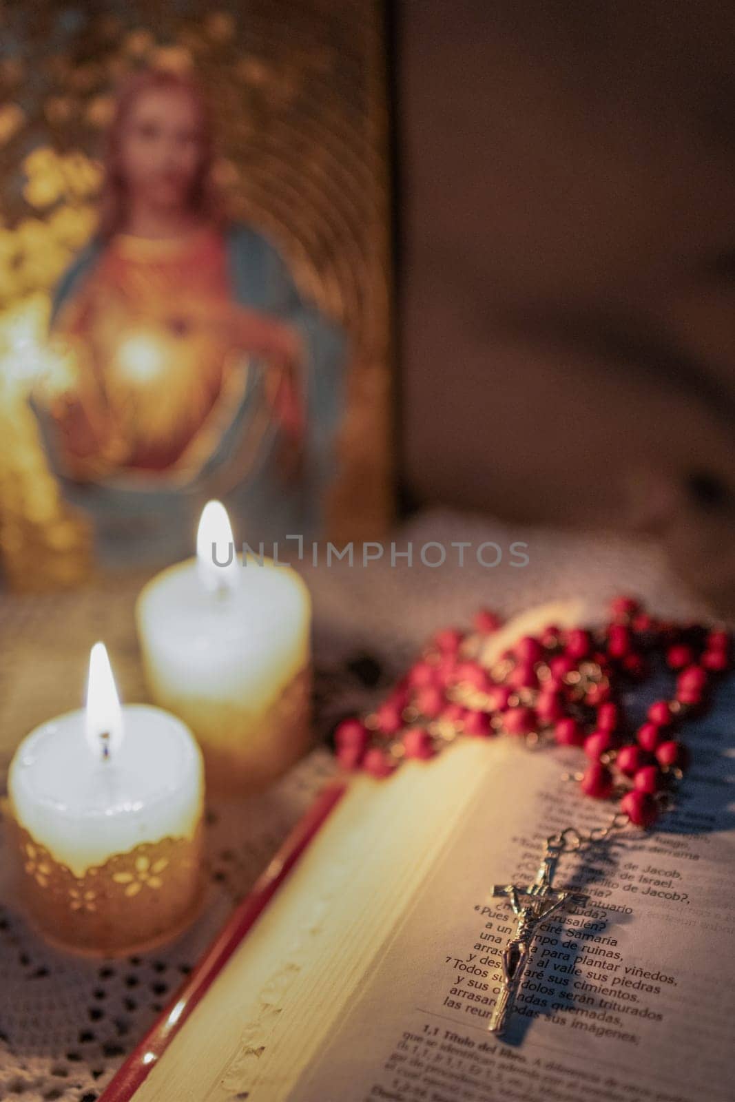 Red rosary on The Bible, illuminated by candlelight.Blurred background by VeroDibe