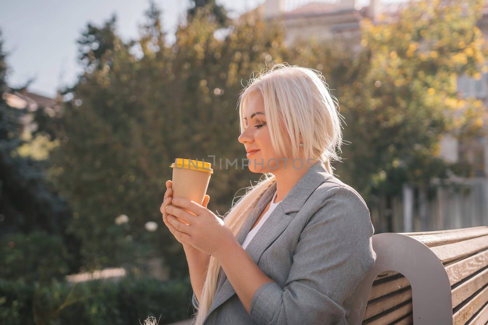 A blonde woman sits on a bench drinking coffee from a yellow cup. She is wearing a gray jacket and has her hair in a ponytail. The scene is peaceful and relaxing