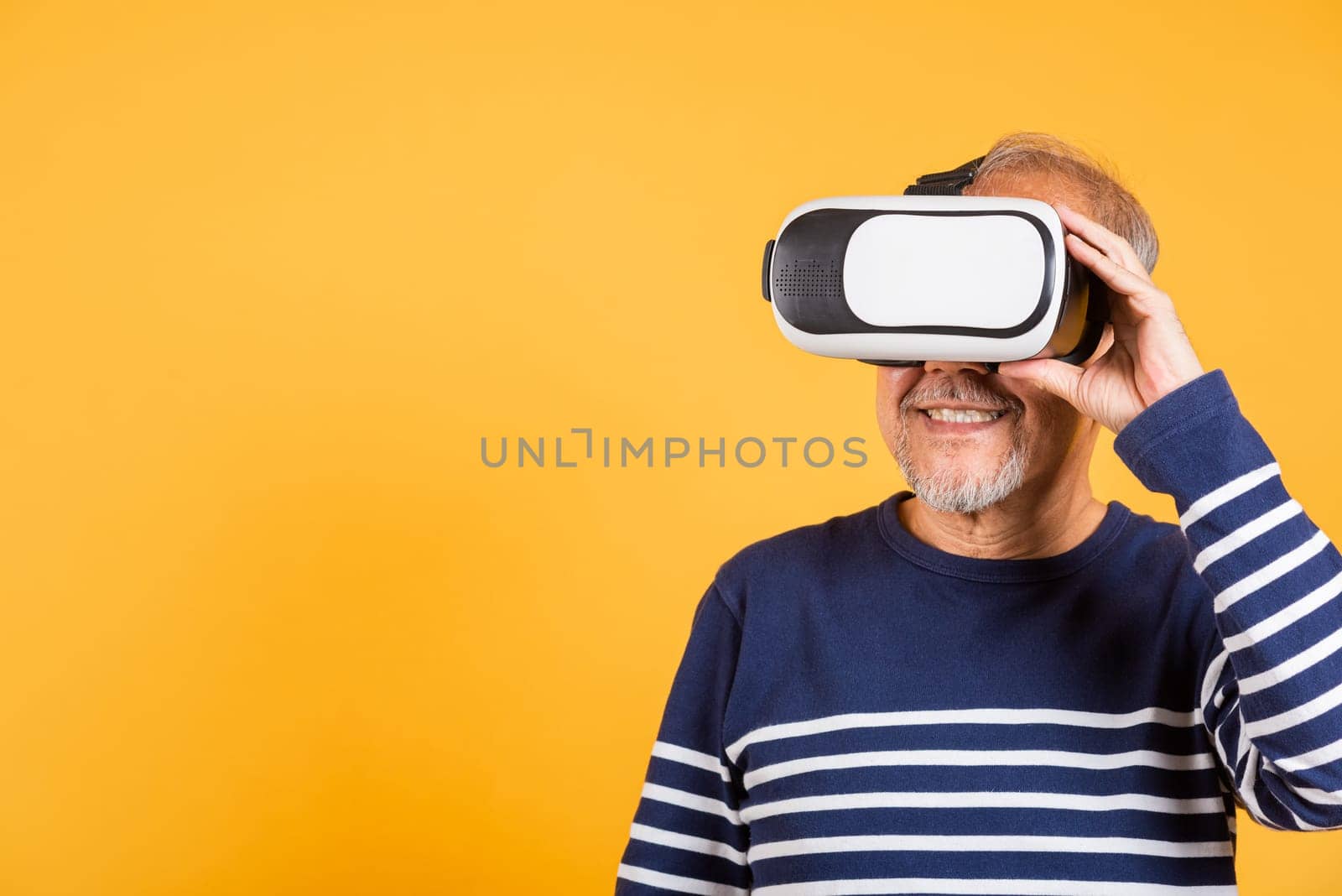 Portrait Asian smiling old man pointing at something in a virtual reality glasses headset studio shot isolated yellow background, Excited happy senior man pensioner using VR by Sorapop