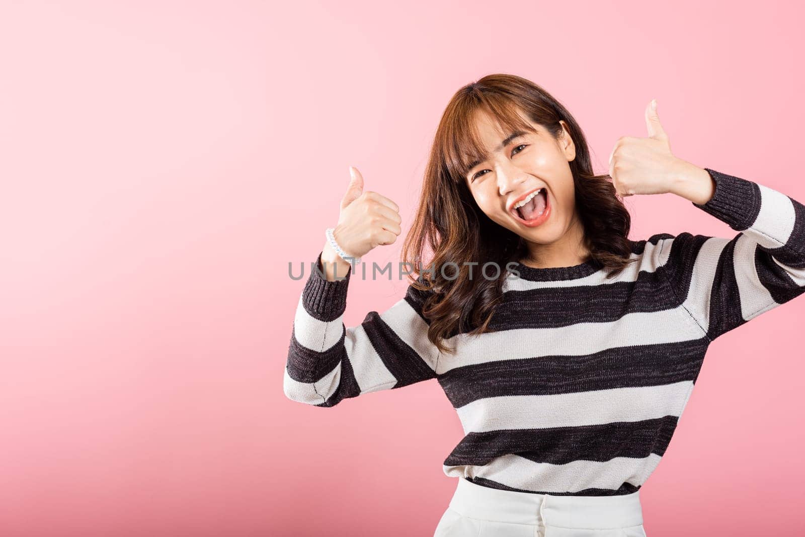 In a pink studio shot, a smiling Asian woman confidently gives a thumbs-up and 'Ok' sign, signifying agreement and success. Her positive gesture radiates joy and satisfaction.