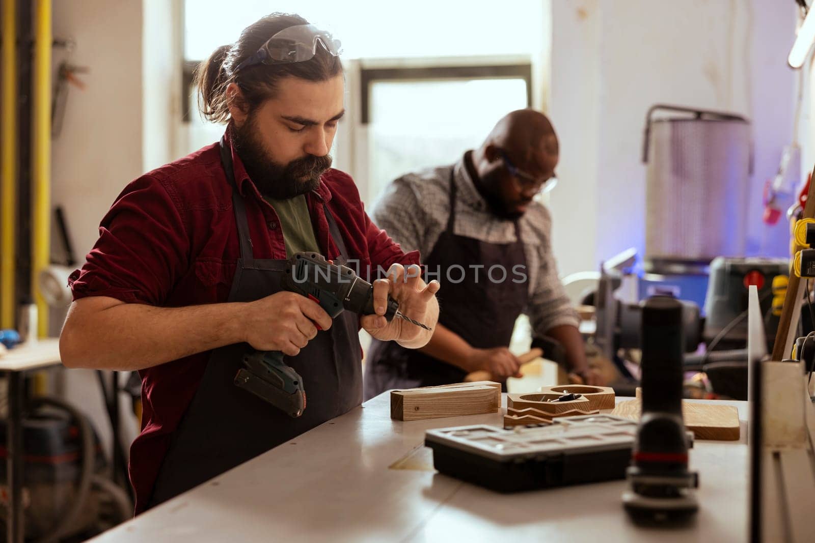 Woodworker next to african american coworker changing power drill head. Carpenter working with BIPOC colleague, preparing to insert screws into wooden surfaces with electric tool