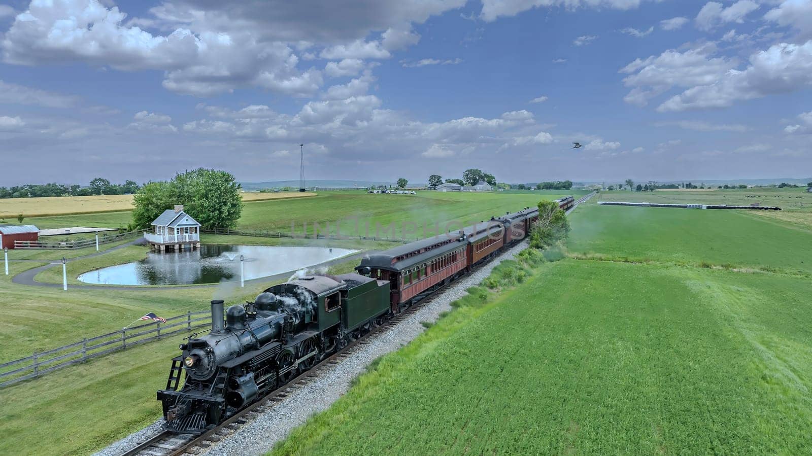A Vintage Steam Train Travels Along A Track Through Lush Green Farmland, Passing A Pond With A Small, Picturesque House, Under A Blue Sky Dotted With Puffy Clouds In A Tranquil Rural Setting.