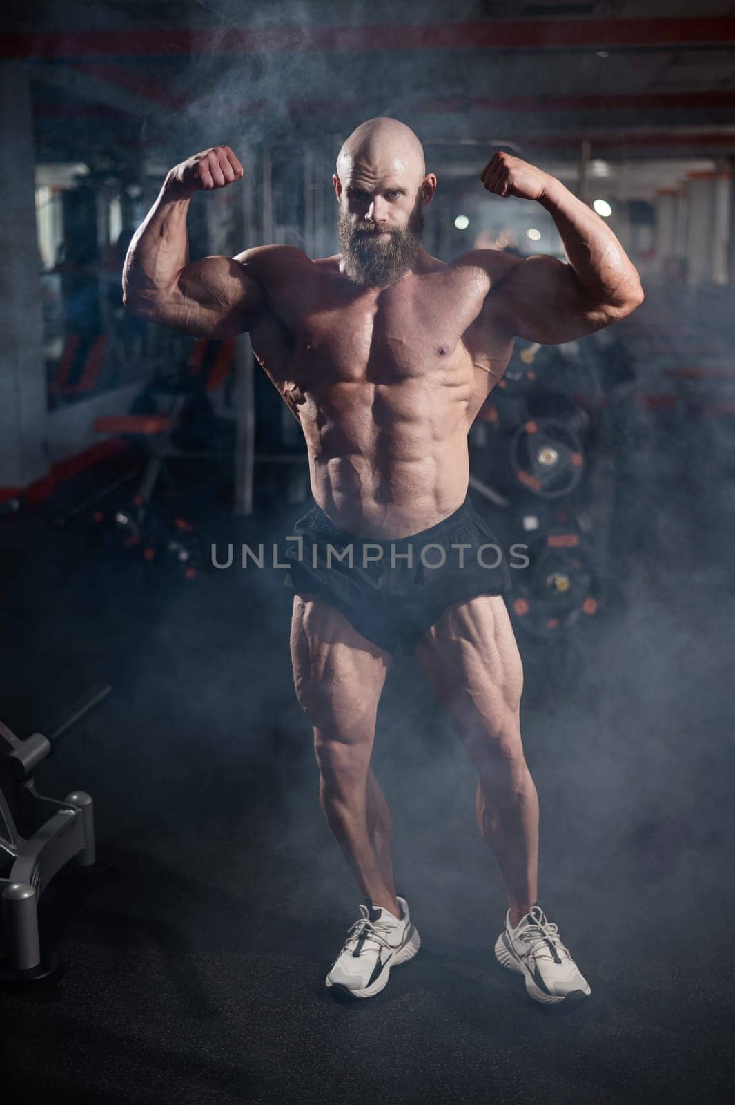 Muscular bald man posing in shorts. Bodybuilder showing off his shape in the gym