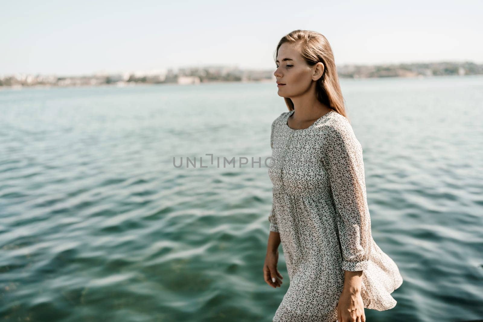 A woman is walking on the beach wearing a dress. The water is calm and the sky is clear