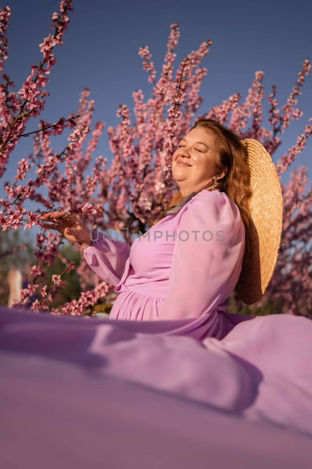 Woman blooming peach orchard. Against the backdrop of a picturesque peach orchard, a woman in a long pink dress and hat enjoys a peaceful walk in the park, surrounded by the beauty of nature