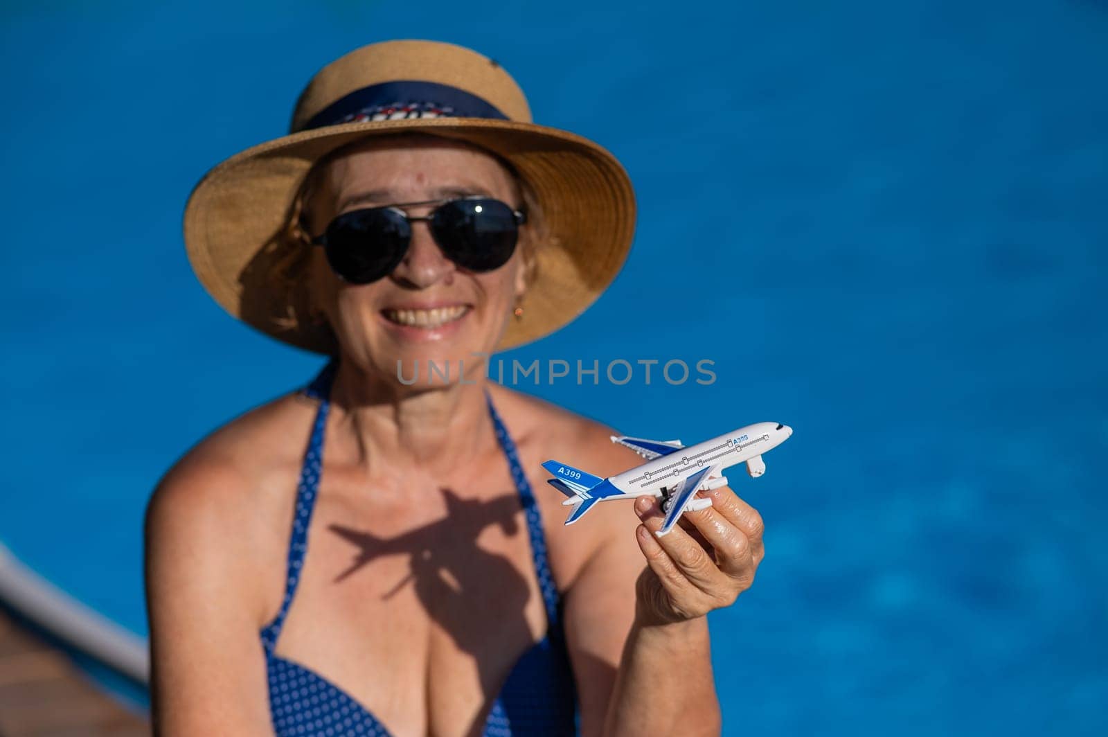 An elderly woman in a hat and sunglasses holds a model airplane while sitting by the pool. Retirement vacation concept