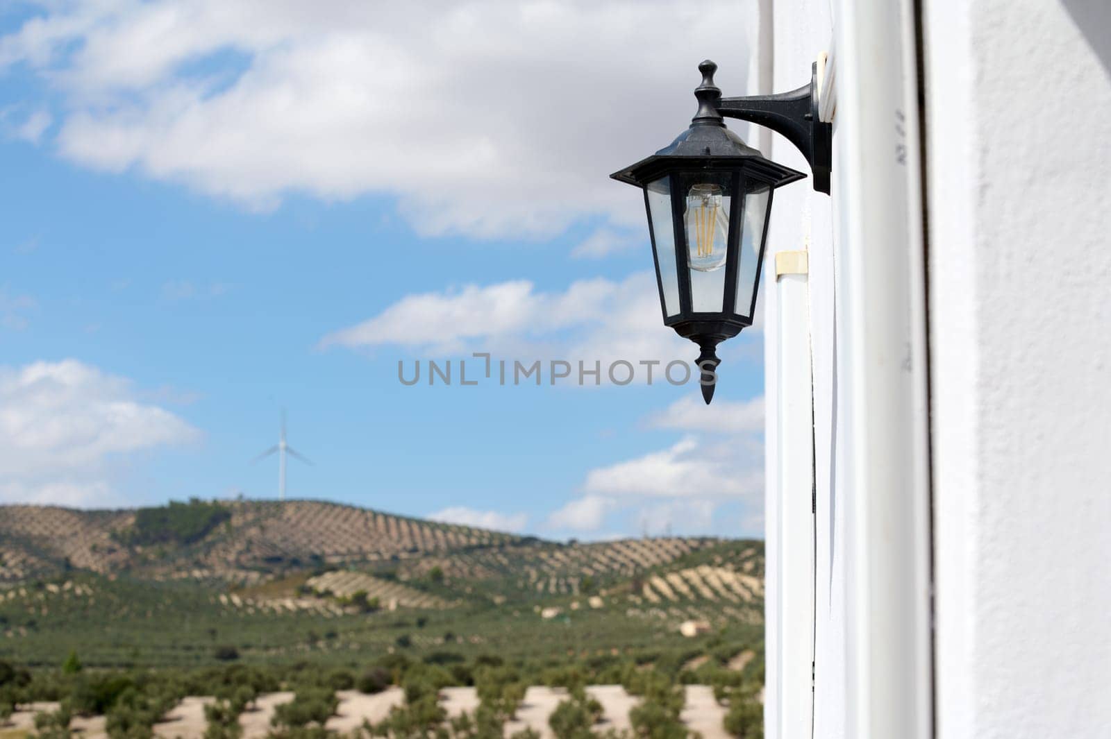 Close-up of a rustic lantern on a white rural house with scenic countryside, rolling hills, and a single wind turbine in the distance under a blue sky.