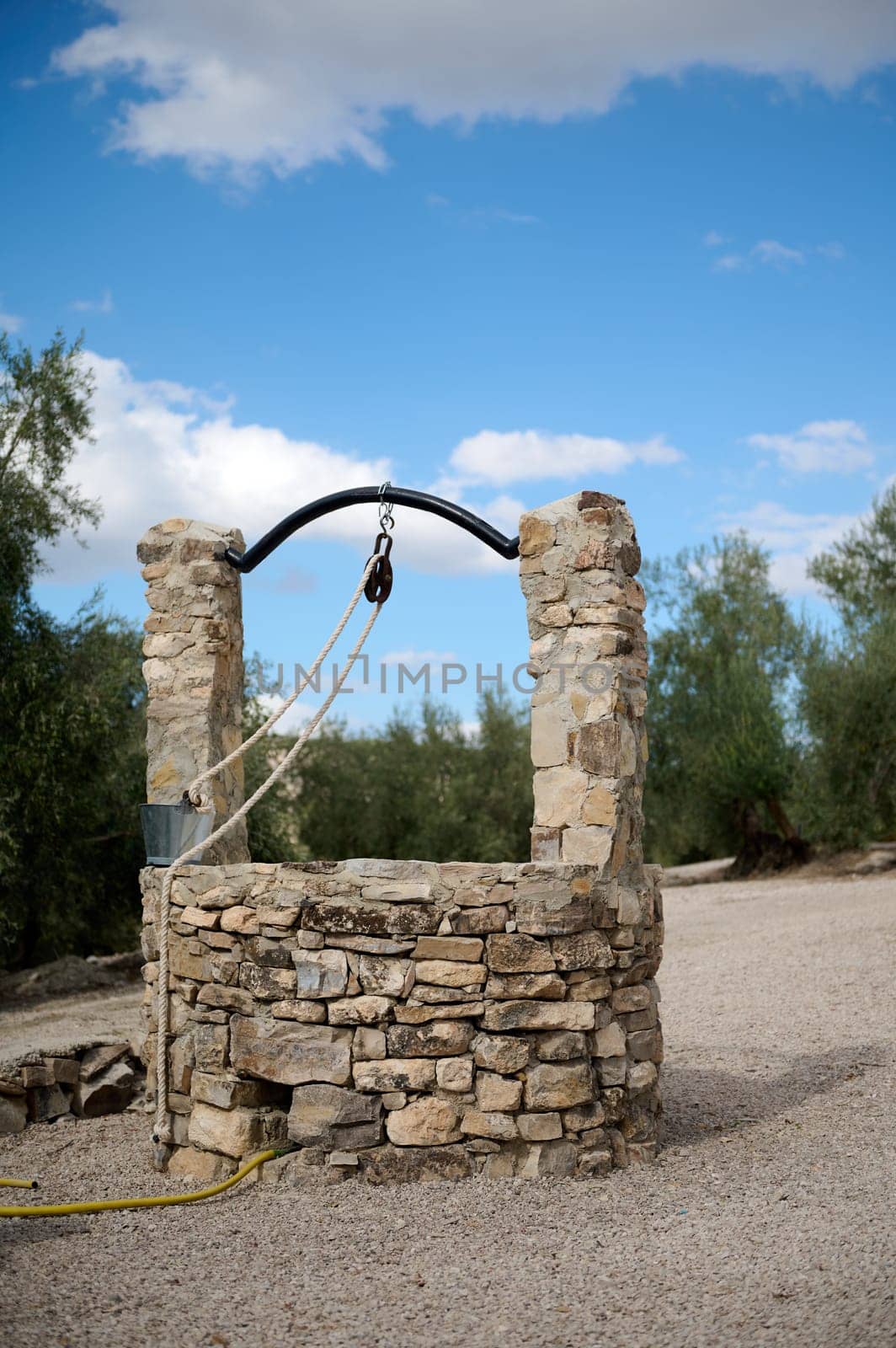 A traditional stone well in a rural landscape, surrounded by trees and under a bright blue sky with clouds. Represents history, countryside life, and simplicity.