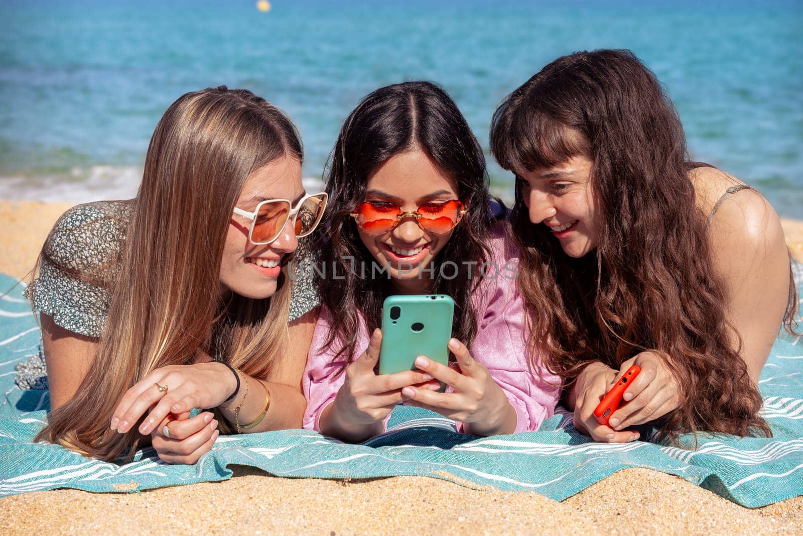 Three smiling friends watching social media while sunbathing on the beach. by mariaphoto3