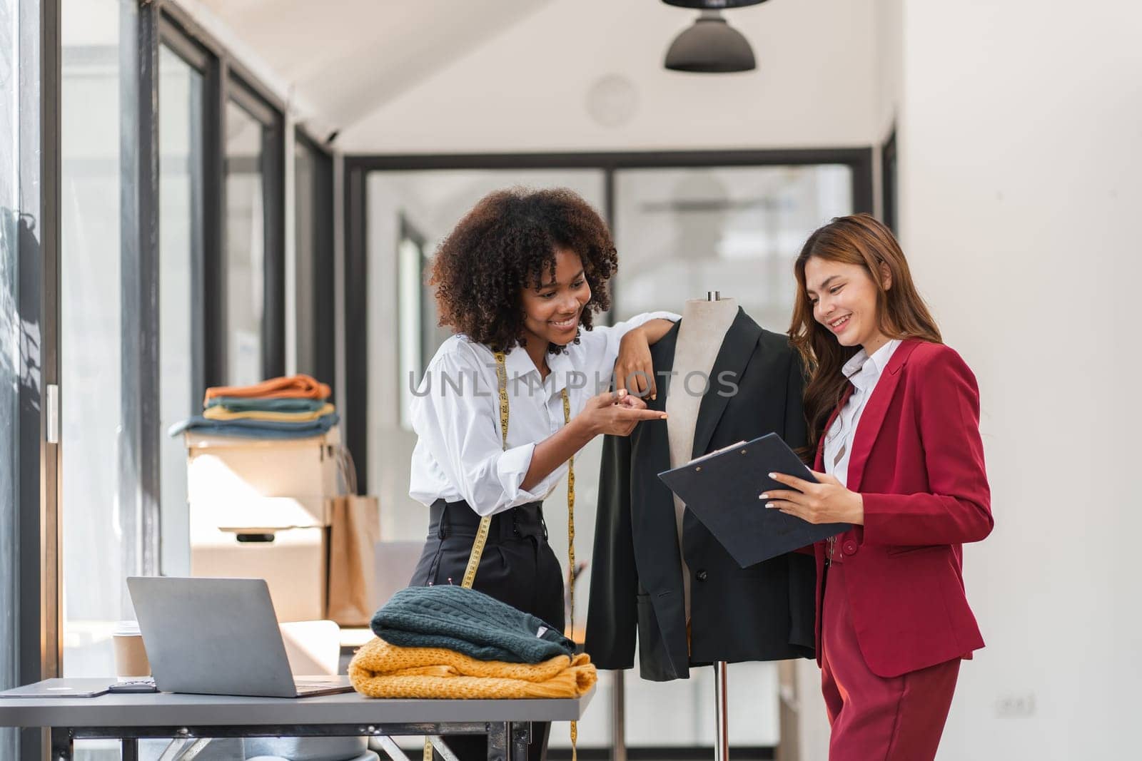 Two female designers working together in a modern studio, creating and designing shirts with a laptop and clothing samples.