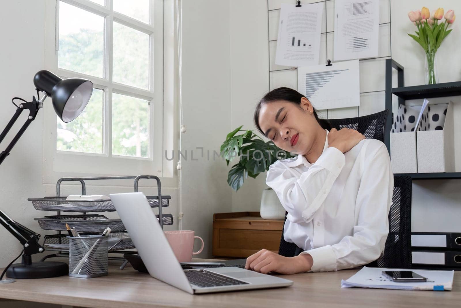 A business professional suffers from neck pain while working at a desk in a modern office, highlighting workplace discomfort and ergonomic issues.