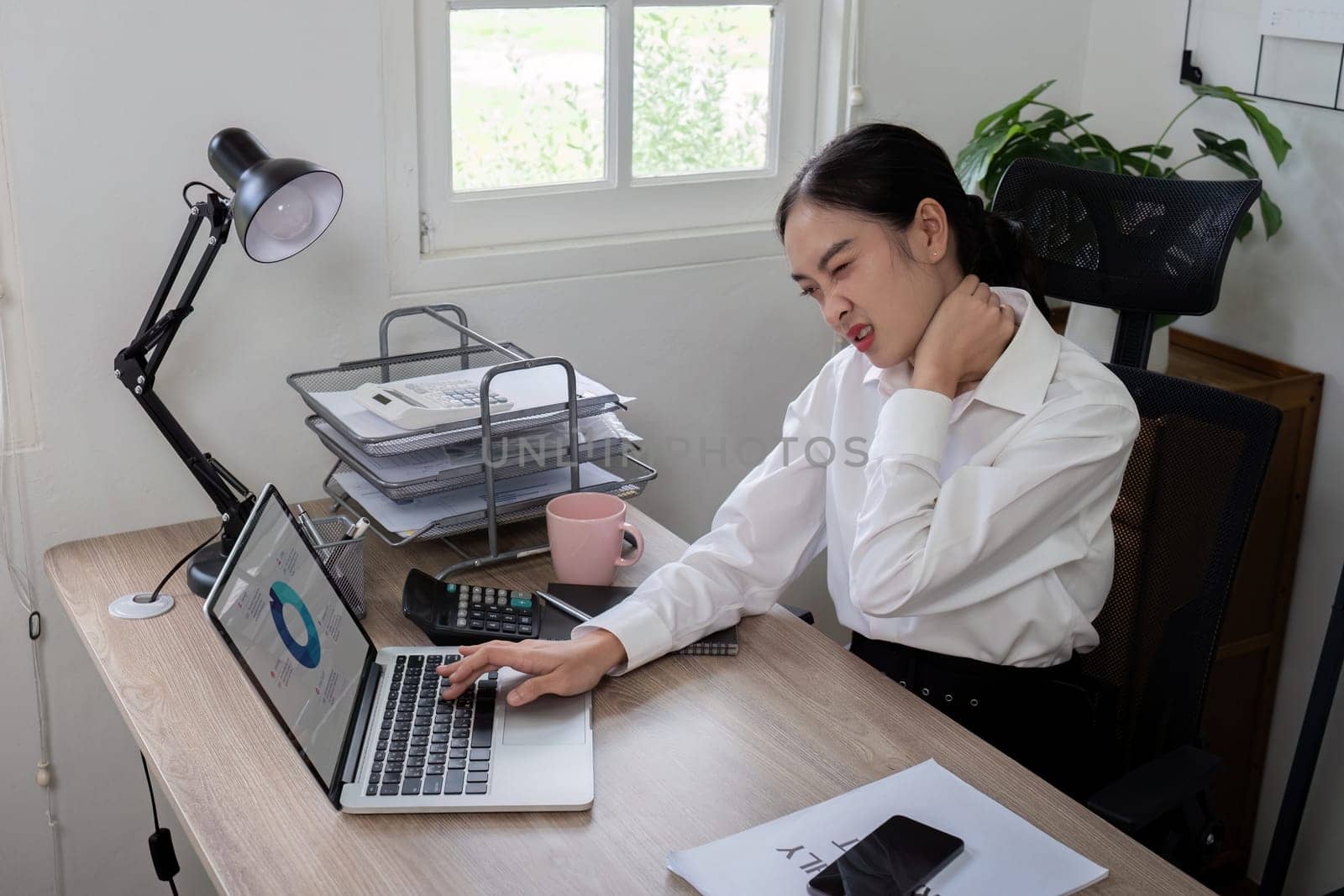 A businesswoman in a modern office setting, experiencing neck pain while working on her laptop. Concept of workplace stress and physical discomfort.