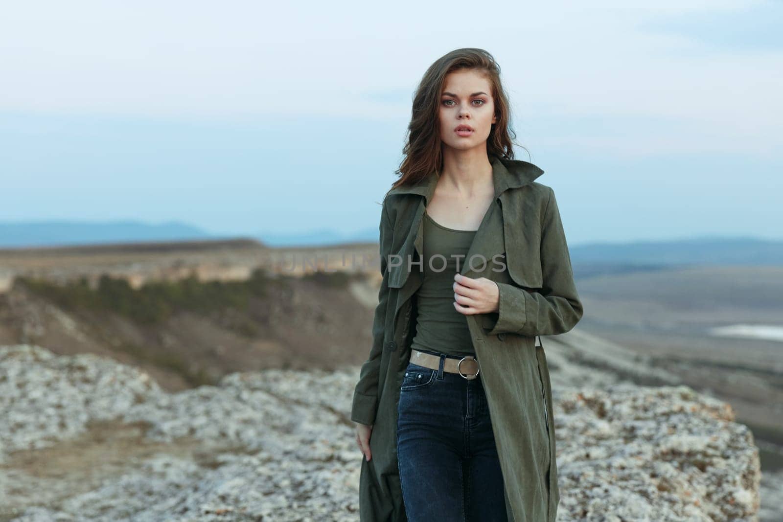 Serene woman in trench coat and jeans standing on rocky hilltop overlooking majestic mountains