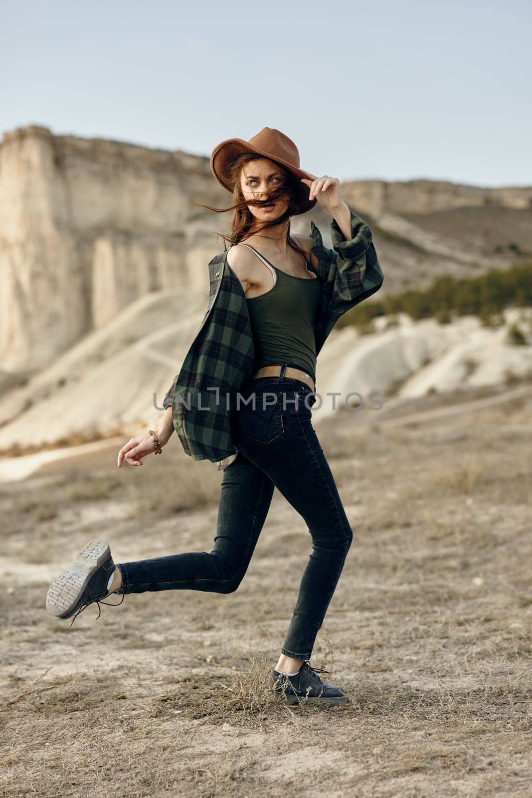 Vibrant young woman joyfully leaping in plaid shirt and hat across desert landscape by Vichizh