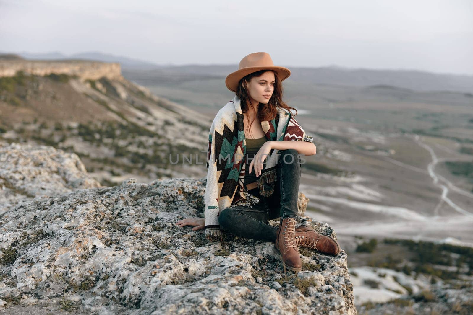 Serene woman in hat and boots sitting on rock in mountainous landscape with majestic mountains in background by Vichizh
