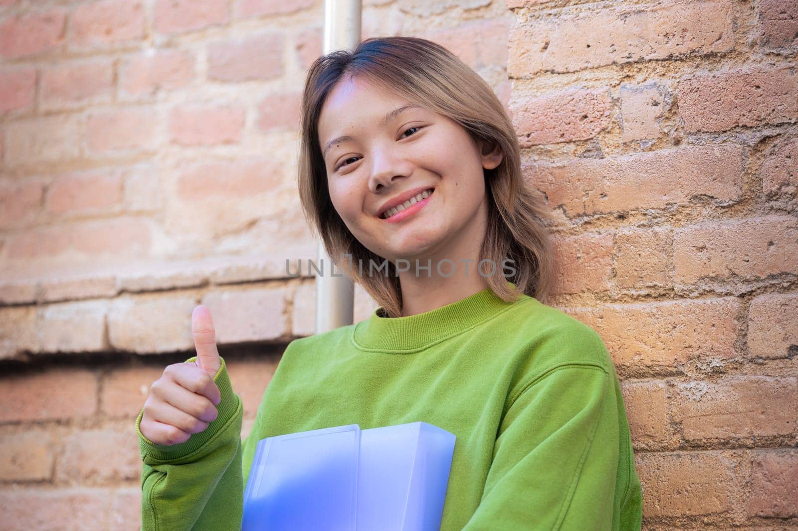 Happy smiling young man holding a notebook and pointing thumb up looking at camera at the university campus