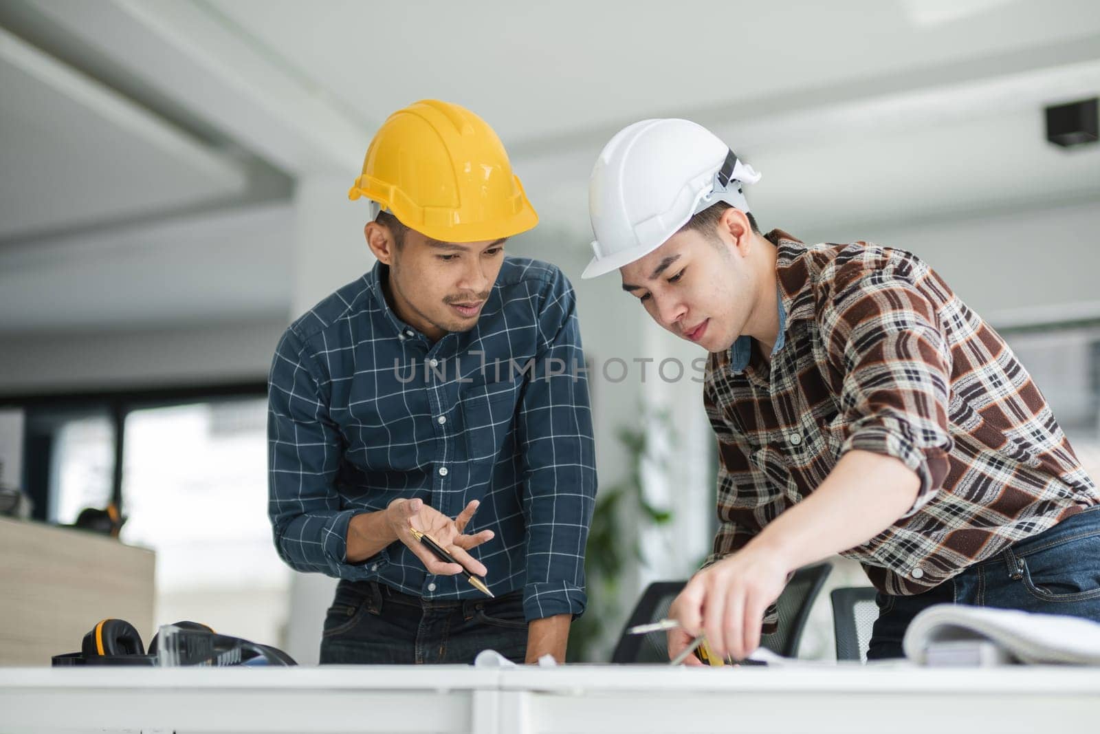 Two male engineers in a modern office, wearing safety helmets and analyzing blueprints, collaborating on project plans.