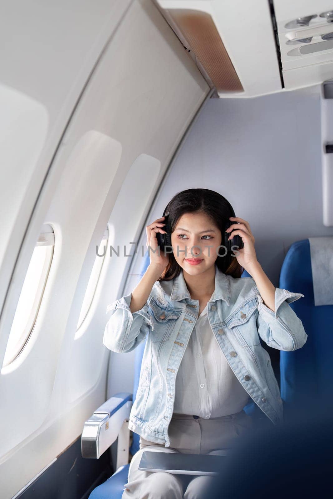 A young female tourist wearing headphones, relaxing in a comfortable airplane seat by the window, enjoying her travel experience.