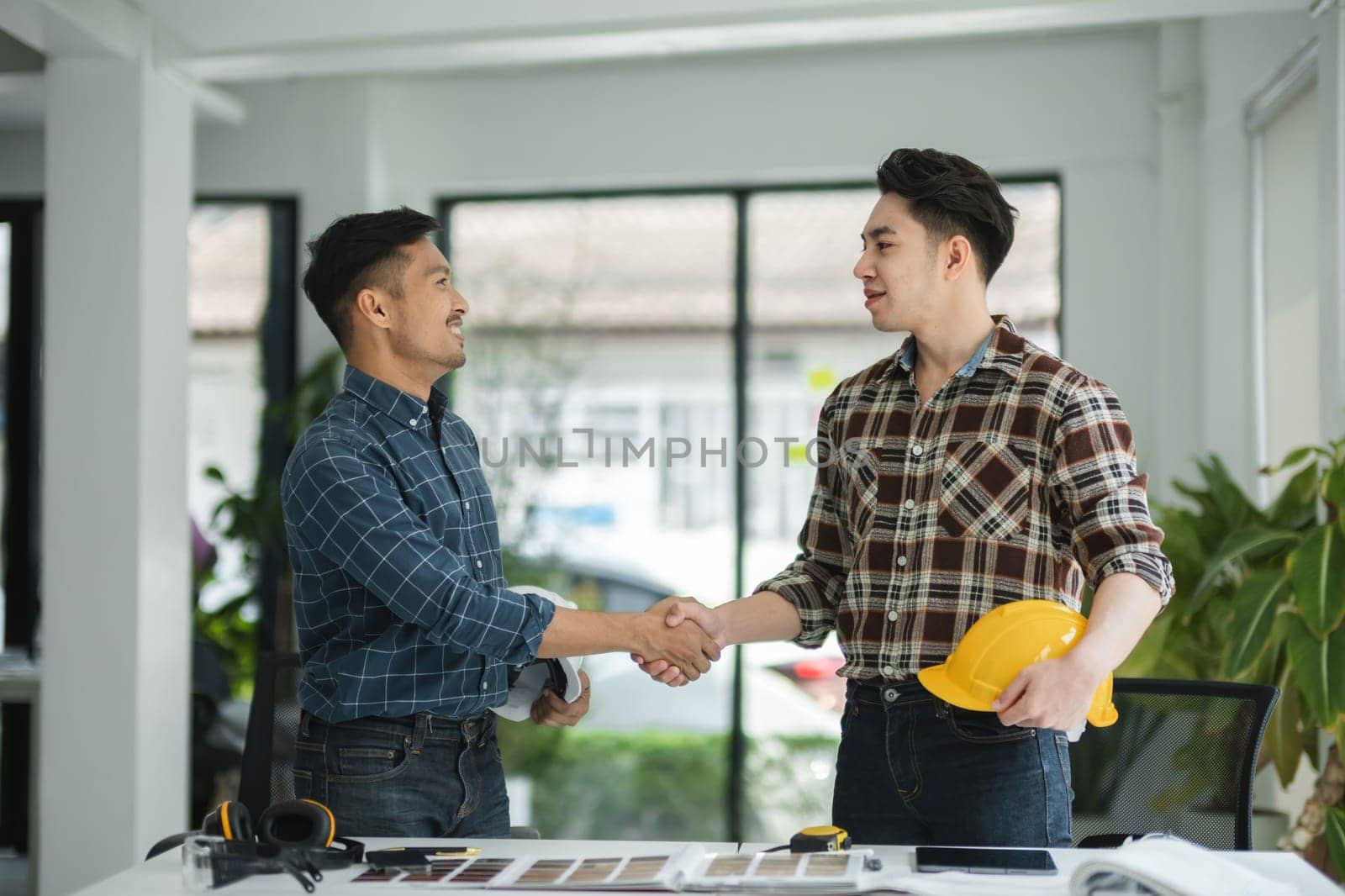Two male engineers in a modern office, shaking hands and discussing project plans, showcasing teamwork and collaboration in a professional environment.