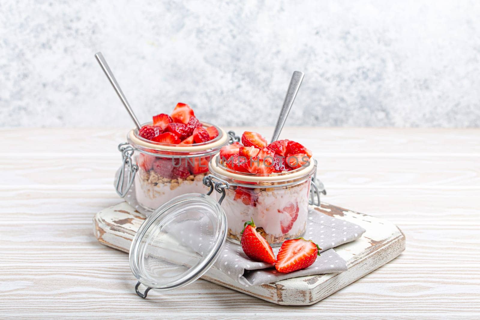 Parfait with Fresh Strawberries, Yoghurt and Crunchy Granola in Transparent Glass Mason Jars on White Rustic Wooden Background Angle View, Healthy Breakfast or Light Summer Fruit Dessert by its_al_dente