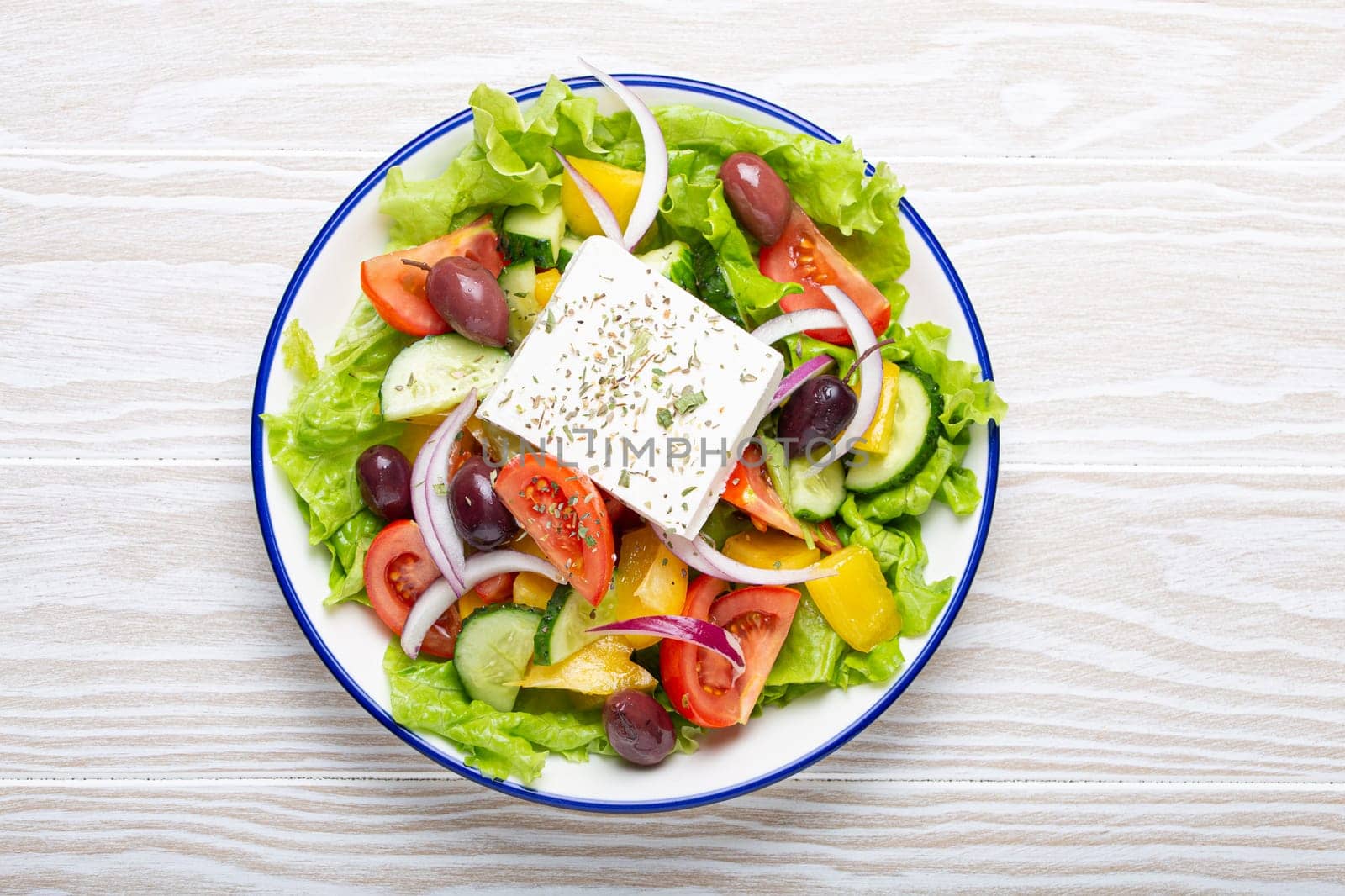 Traditional Greek Salad with Feta Cheese, Tomatoes, Bell Pepper, Cucumbers, Olives, Herbs in white ceramic bowl on White rustic wooden table background from above, Cuisine of Greece