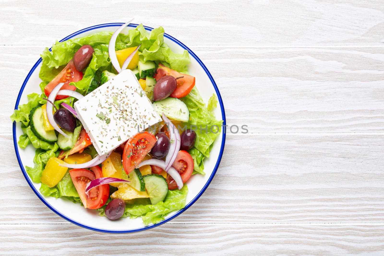 Traditional Greek Salad with Feta Cheese, Tomatoes, Bell Pepper, Cucumbers, Olives, Herbs in white ceramic bowl on White rustic wooden table background from above, Cuisine of Greece. Copy space